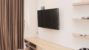 A modern room with a flat-screen TV mounted on the wall, a wooden bench, and a small shelf with a decorative item. - Minimalist And Cozy Studio At Vasanta Innopark Apartment (Cikarang)