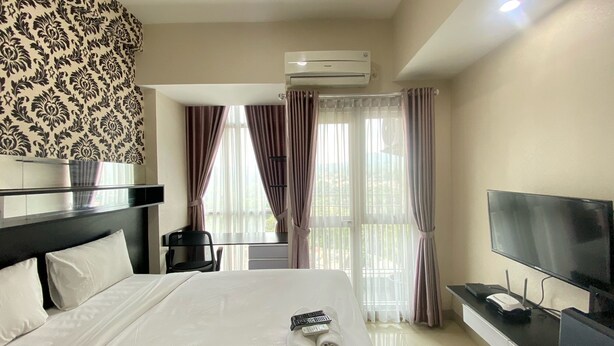 A hotel room with a bed, a TV, an air conditioner, and a view of the outdoors through the window. - Affordable Studio Room At Taman Melati Jatinangor Apartment (Hegarmanah)