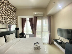 A hotel room with a bed, a TV, an air conditioner, and a view of the outdoors through the window. - Affordable Studio Room At Taman Melati Jatinangor Apartment (Hegarmanah)