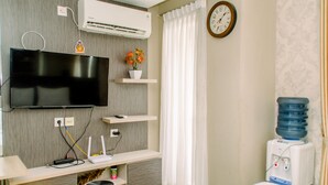 A hotel room with a flat-screen TV, a desk with a chair, a water dispenser, and a clock on the wall. - Minimalist And Comfy Studio At Bintaro Icon Apartment (South Tangerang)
