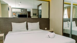 A neatly made bed with white linens and two rolled towels, a wooden headboard, and a large mirror reflecting the room's interior. - Minimalist And Comfy Studio At Bintaro Icon Apartment (South Tangerang)