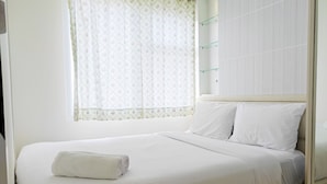 Een net opgemaakt bed met witte lakens en een opgerolde handdoek, een raam met bloemen gordijnen, en een wandplank met glazen planken.