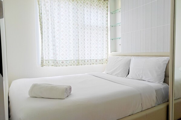 A neatly made bed with white linens and a rolled towel, a window with floral curtains, and a wall-mounted shelf with glass shelves.