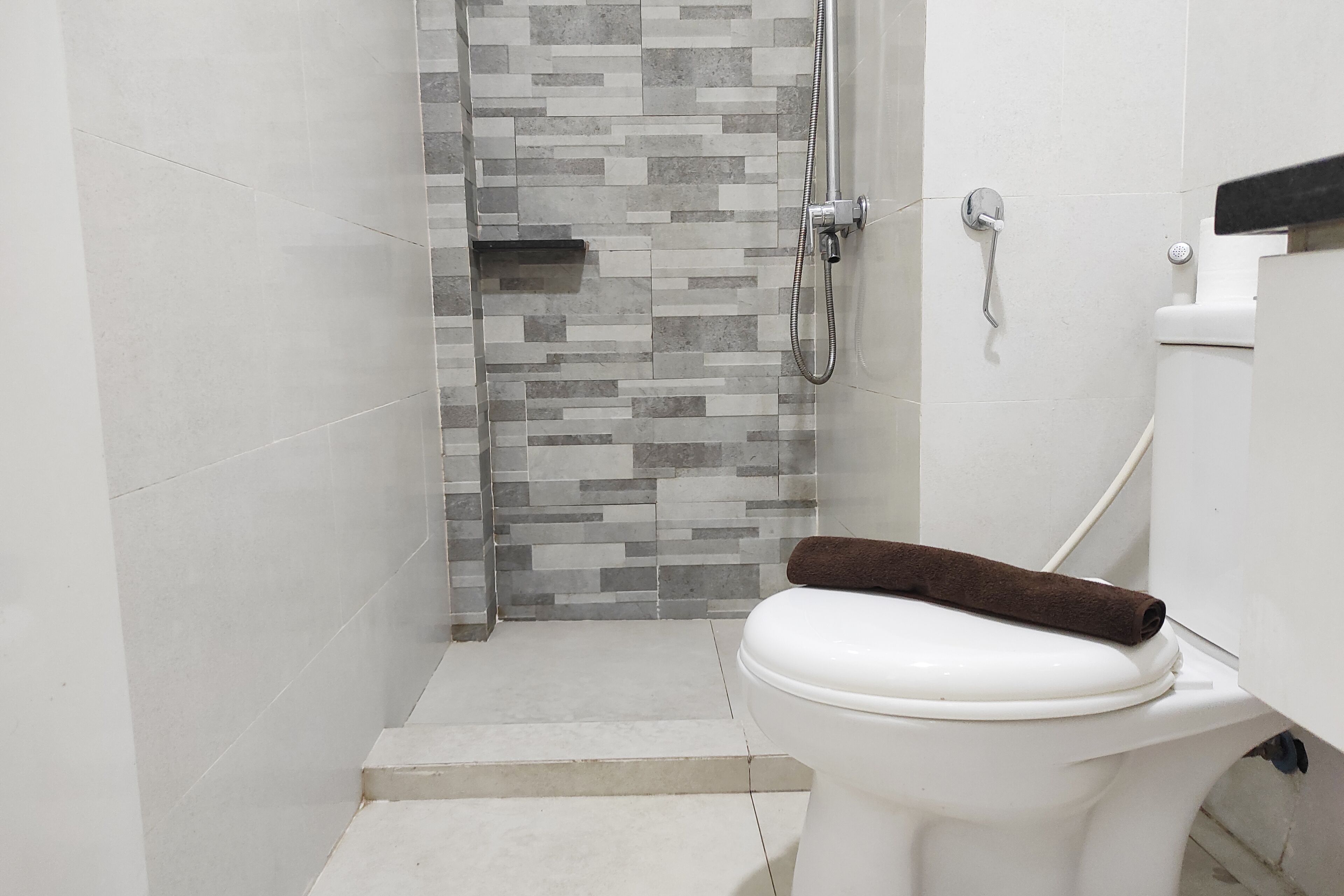 A bathroom with a white toilet, a shower with a tiled wall, and a towel on the toilet.