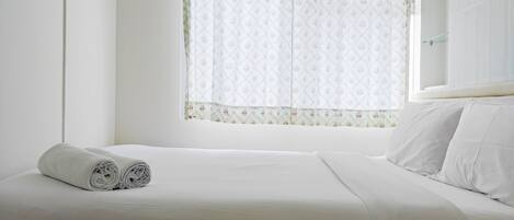 Una cama bien hecha con sábanas blancas y una toalla enrollada encima, una ventana con cortinas florales y una pared blanca.