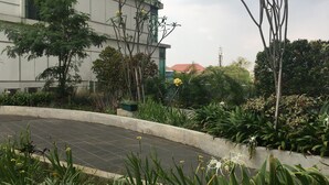 A landscaped area with a curved pathway, various plants, and a building with large windows in the background. - Spacious Studio Room At Tamansari La Grande Apartment Near Bip (Bandung)