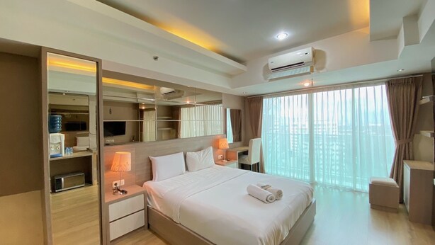 A modern hotel room with a large bed, a built-in wardrobe, a desk with a lamp, and a large window with curtains. - Spacious Studio Room At Tamansari La Grande Apartment Near Bip (Bandung)
