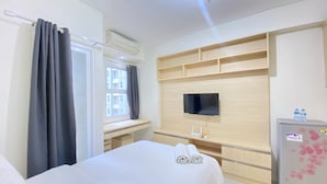 Una camera d'albergo con un letto, una TV a muro, una scrivania con un computer, una finestra con tende e un frigorifero con bottiglie d'acqua sopra.