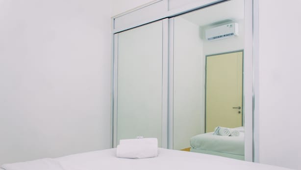 A hotel room with a bed, a white towel on the bed, a sliding door, and an air conditioning unit on the wall. - Warm And Minimalist 3Br Apartment At M-Town Residence (Gading Serpong)
