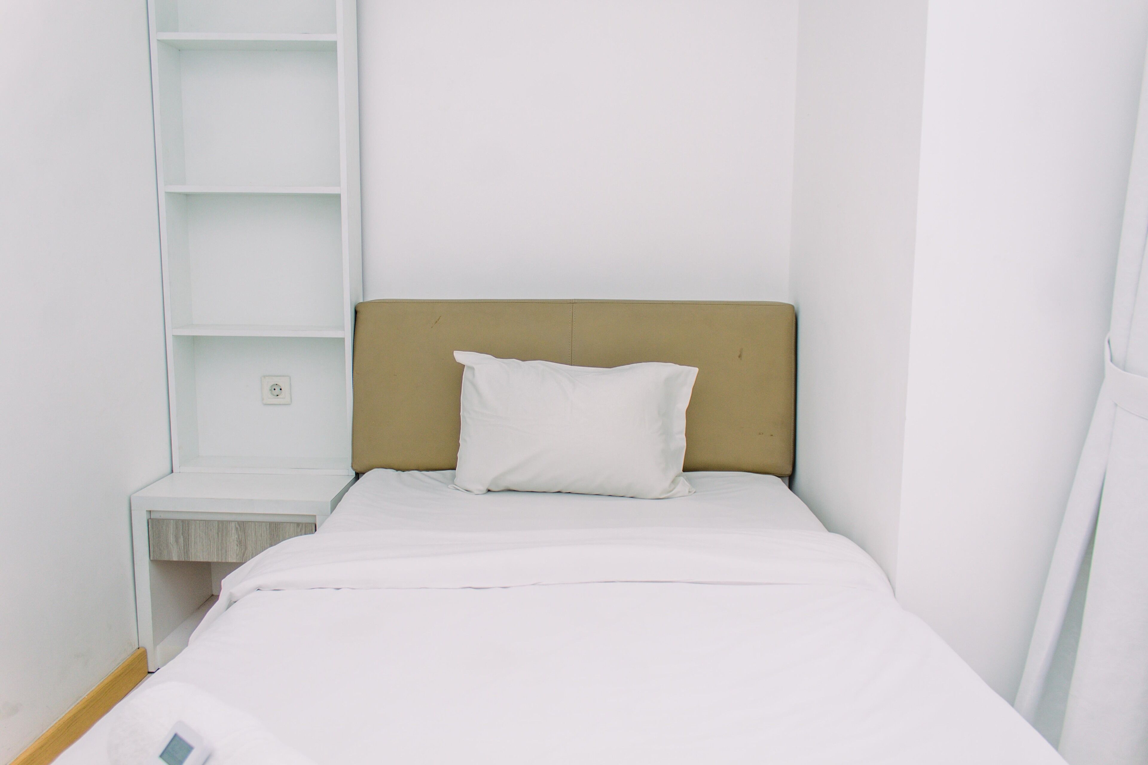A small, neatly made bed with a white pillow and a beige headboard in a minimalistic room with white walls and a white nightstand.