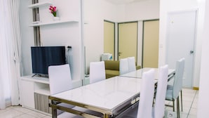 A modern dining area with a glass table, white chairs, a flat-screen TV, and a sofa. - Warm And Minimalist 3Br Apartment At M-Town Residence (Gading Serpong)