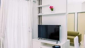 A modern living room with a flat-screen TV on a white stand, a small white shelf with a flower, and a green sofa. - Warm And Minimalist 3Br Apartment At M-Town Residence (Gading Serpong)