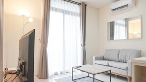 A modern living room with a flat-screen TV, a grey sofa, a wooden coffee table, and a large window with sheer curtains. - Nice And Comfort 2Br At Pejaten Park Apartment (Jakarta)