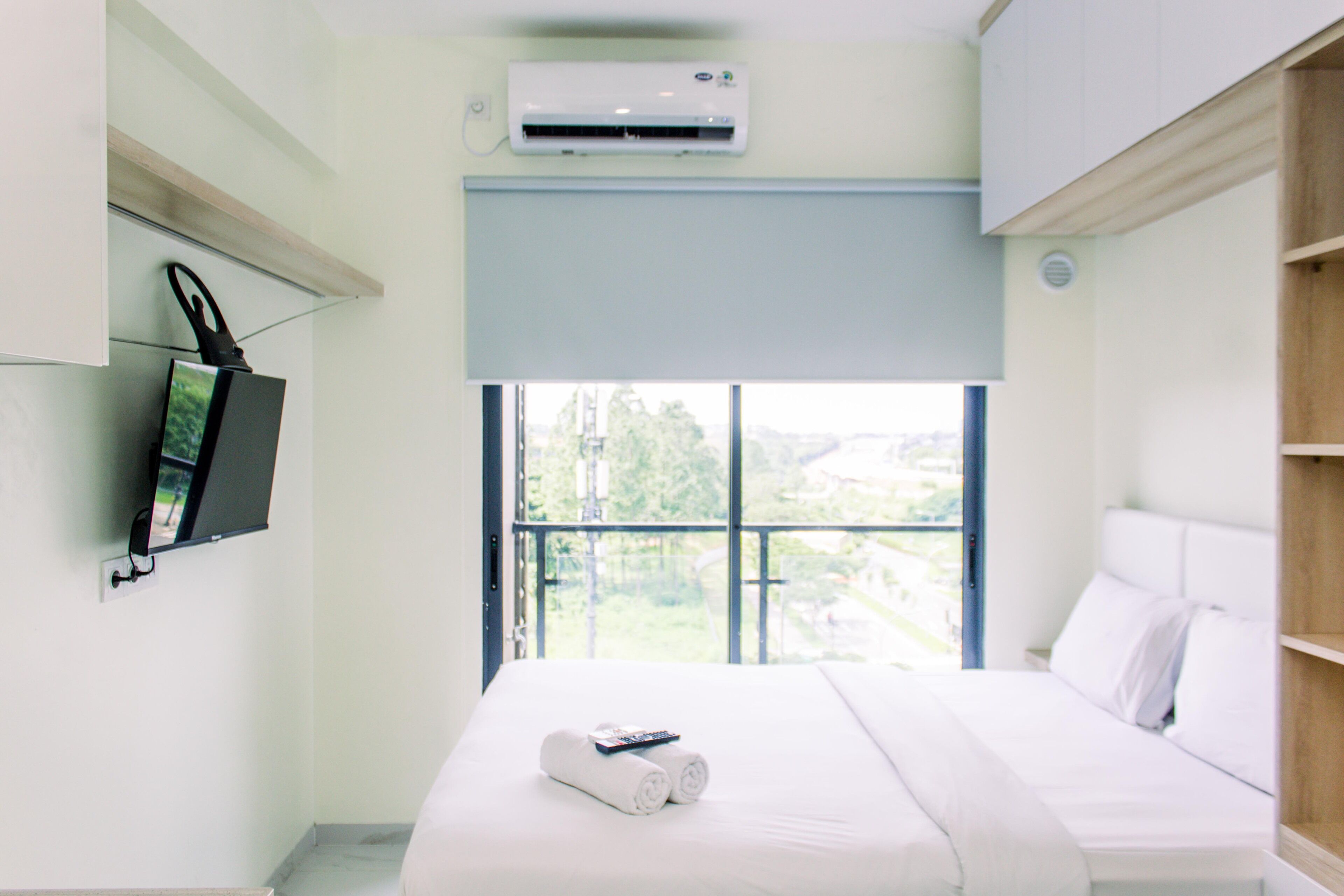 A hotel room with a bed, a TV mounted on the wall, and a window with a view of greenery.