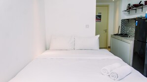 Uma cama pequena e bem arrumada com lençóis brancos e duas toalhas enroladas, uma cozinha compacta com geladeira e fogão, e uma porta que dá para outro cômodo.