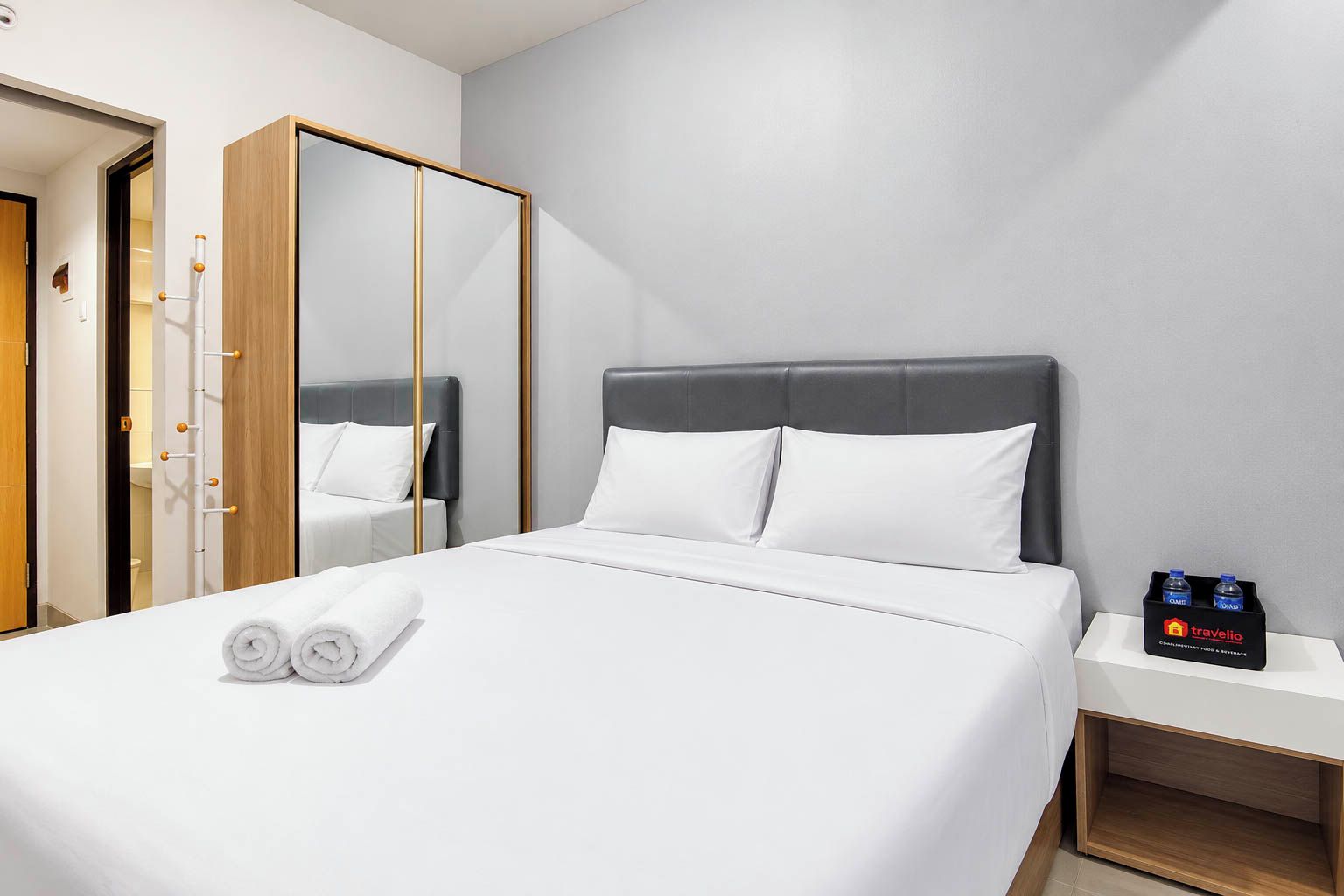 A hotel room with a bed, a mirror, a nightstand with bottled water, and a wooden wardrobe.