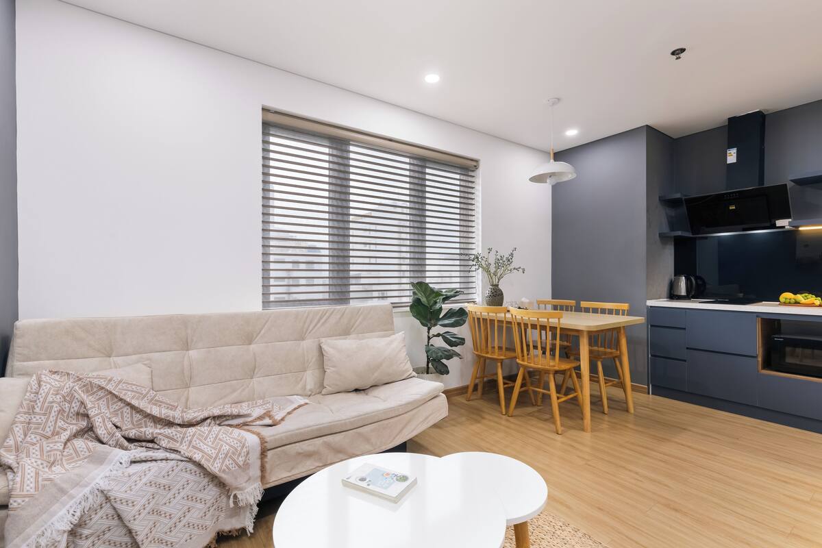 Uma sala de estar moderna com um sofá bege, uma mesa de jantar com cadeiras, uma mesa de centro branca e uma área de cozinha com micro-ondas e fogão.