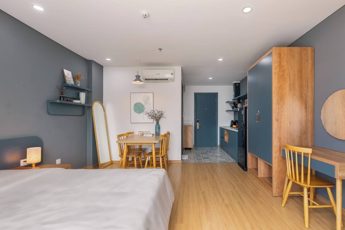 Quarto de hotel moderno com cama, mesa de jantar de madeira, cadeiras, pequena cozinha e um banheiro visível através de uma porta aberta.