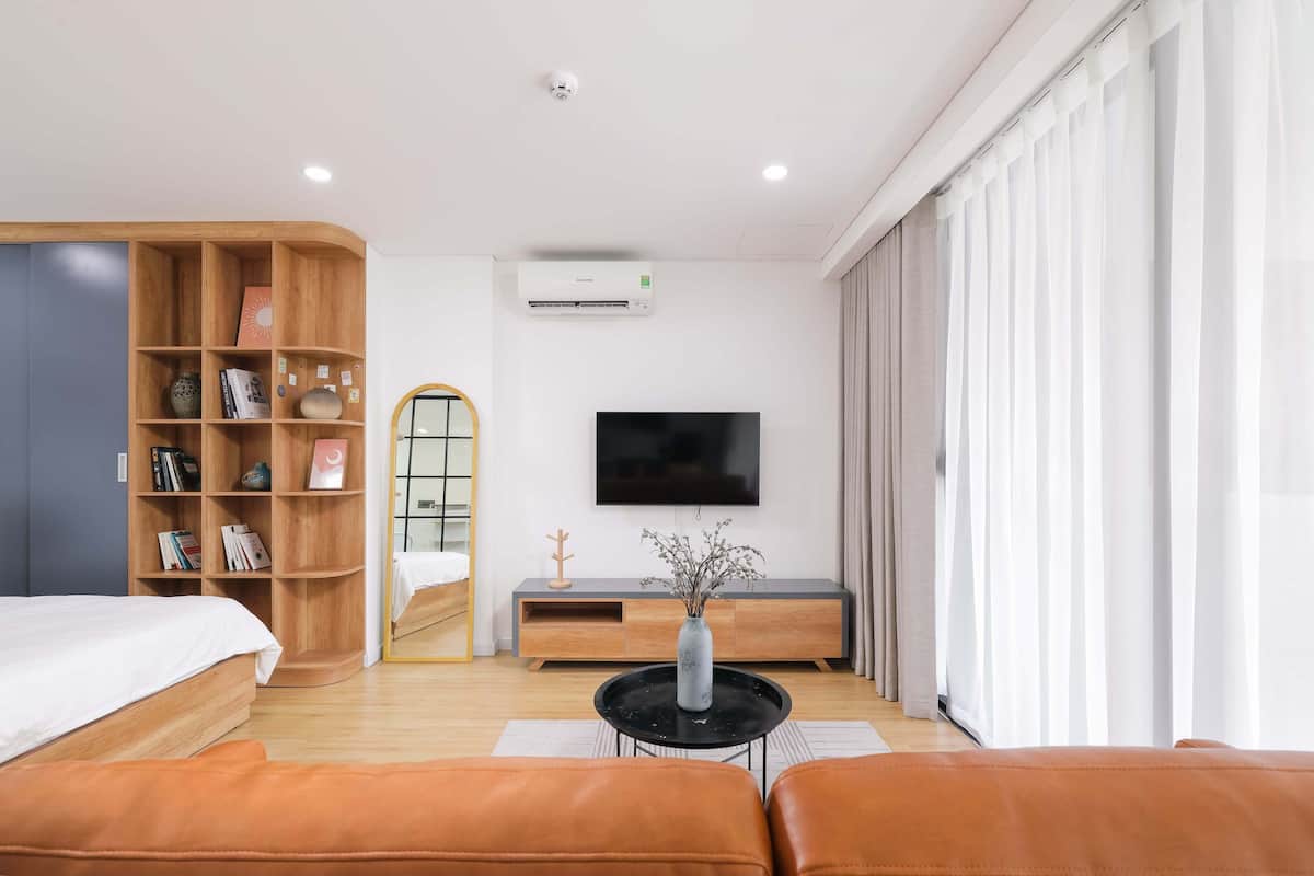 Quarto de hotel moderno com cama, estante de madeira, TV de tela plana, pequena mesa com um vaso e uma janela grande com cortinas.