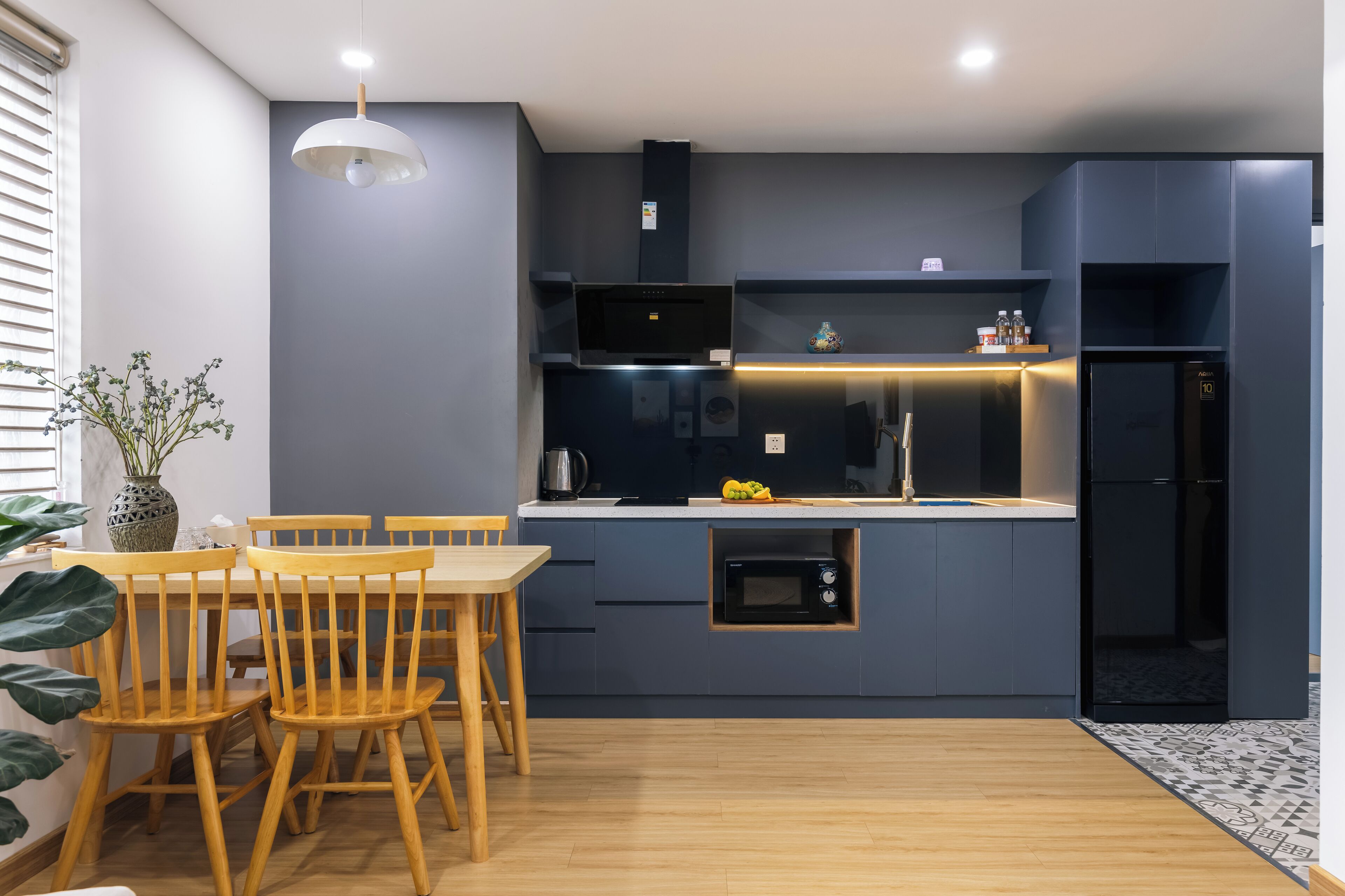 A modern kitchen with dark cabinetry, a built-in microwave, and a refrigerator. There is a dining table with chairs and a pendant light above it.