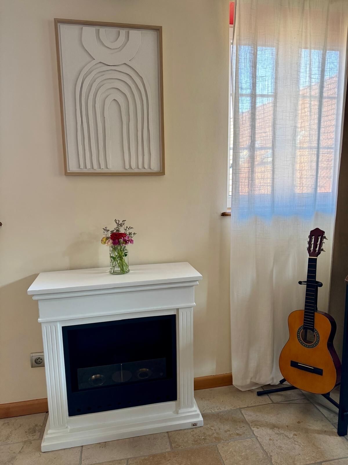 A room with a white fireplace, a guitar leaning against the wall, and a window with sheer curtains.