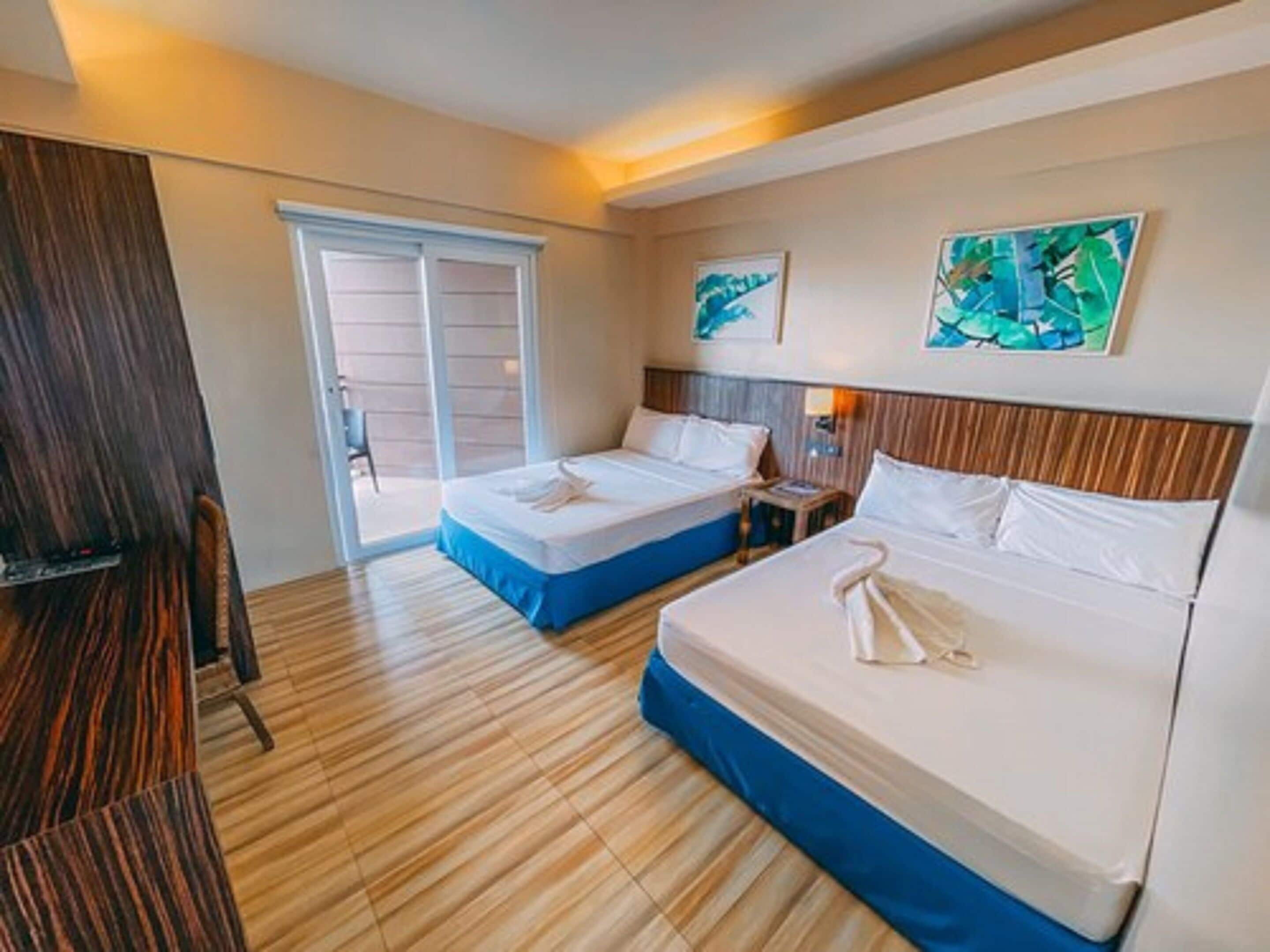 A hotel room with two beds, a wooden floor, and a sliding glass door leading to an outdoor area.
