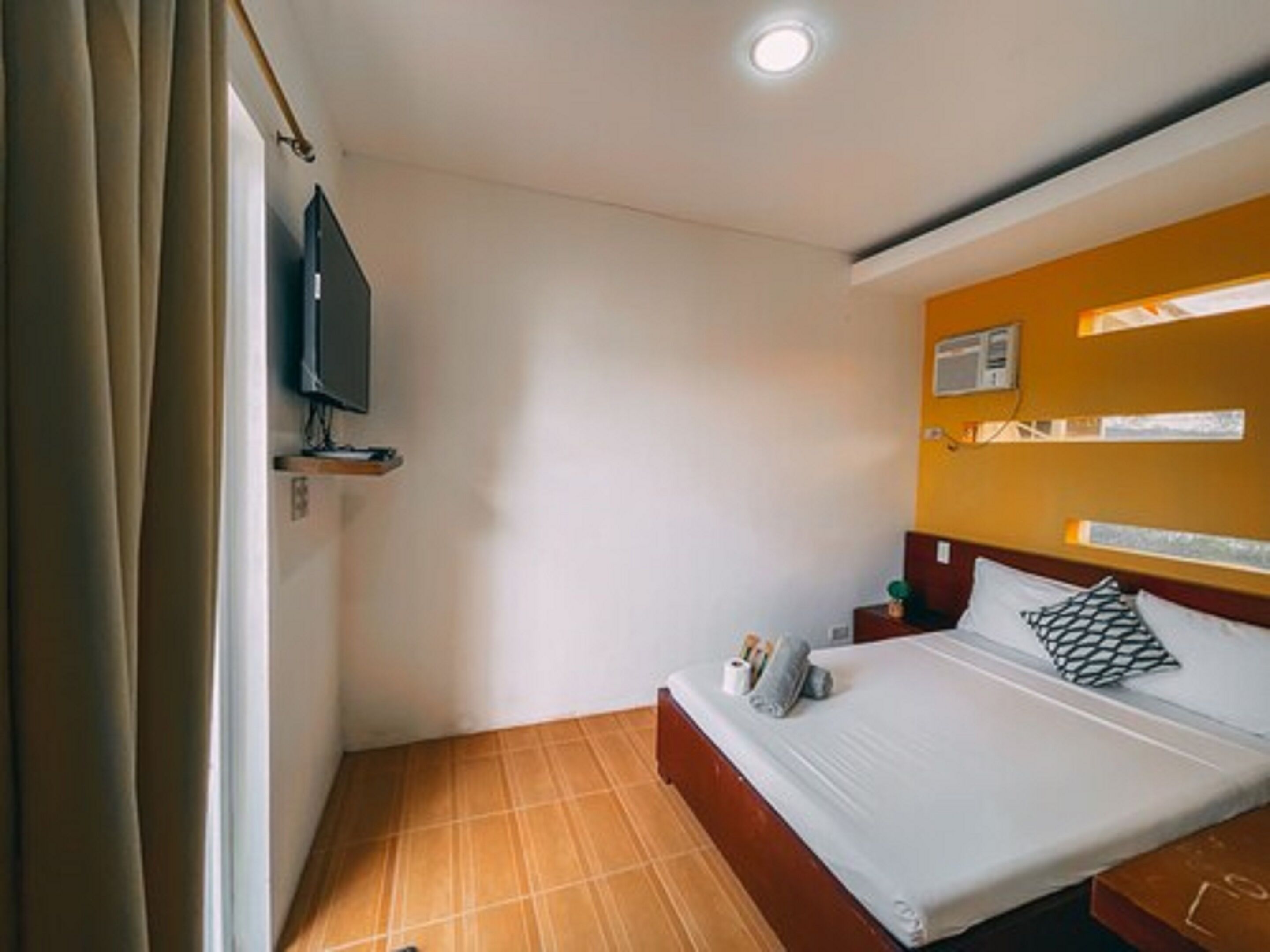 A hotel room with a bed, a TV mounted on the wall, a small bedside table, and a window with curtains.