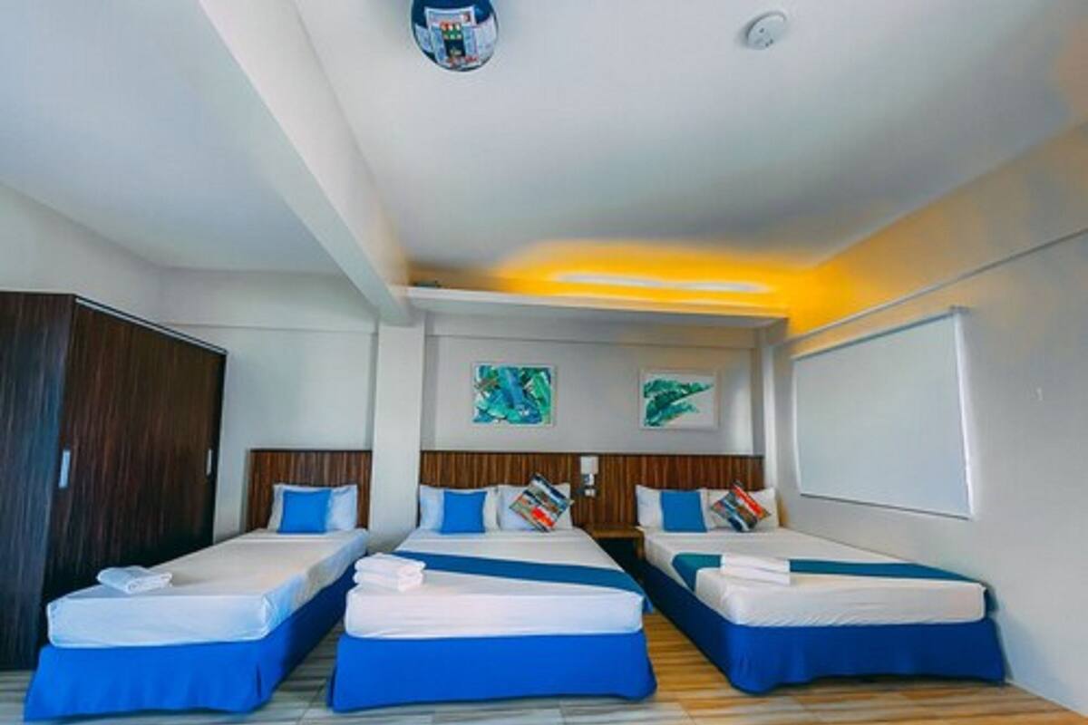 A hotel room with three single beds, each with a blue bedspread and white pillows. There is a wooden wardrobe on the left, a ceiling light, and two wall-mounted artworks.
