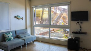 Un salon avec un canapé gris, une grande fenêtre donnant sur un escalier et une zone extérieure, et un téléviseur à écran plat fixé au mur.