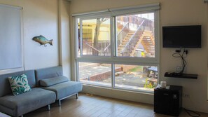 A living room with a grey sofa, a large window with a view of a staircase and outdoor area, and a flat-screen TV mounted on the wall.