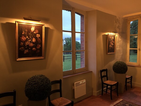 Een gezellig verlichte gang met een groot bloemschilderij, een ronde potplant en twee stoelen.