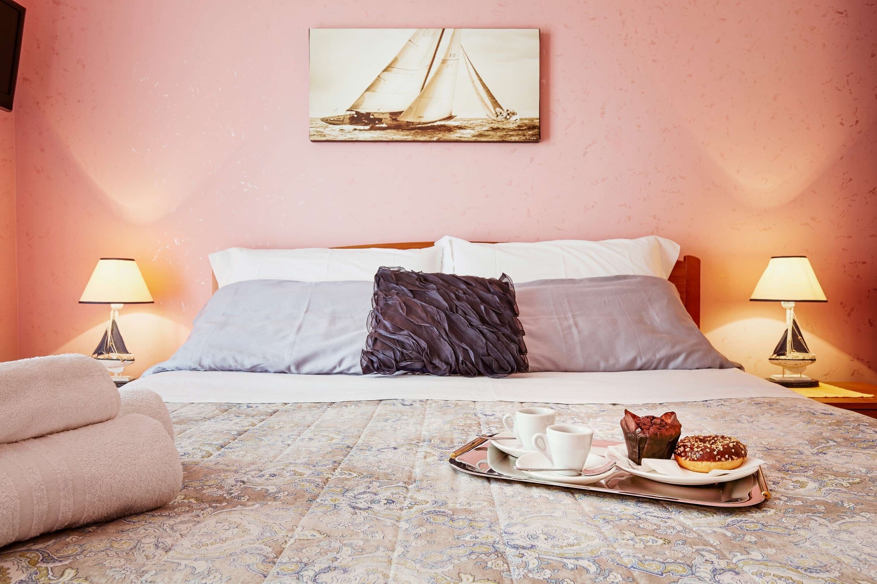 Een hotelkamer met een bed, twee bedlampen, een dienblad met gebak en een schilderij van zeilboten aan de muur.