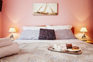 A hotel room with a bed, two bedside lamps, a tray with pastries, and a painting of sailboats on the wall.