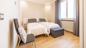A bedroom with two beds, a chair, a small table, and a window with curtains. - Holiday Apartment 'New Casa Ilaria' near Center with Garden & Wi-Fi (Torri del Benaco)