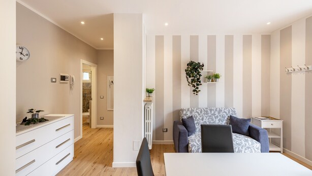 A modern room with a dining table, a sofa, a white dresser, a clock, and a radiator. - Holiday Apartment 'New Casa Ilaria' near Center with Garden & Wi-Fi (Torri del Benaco)