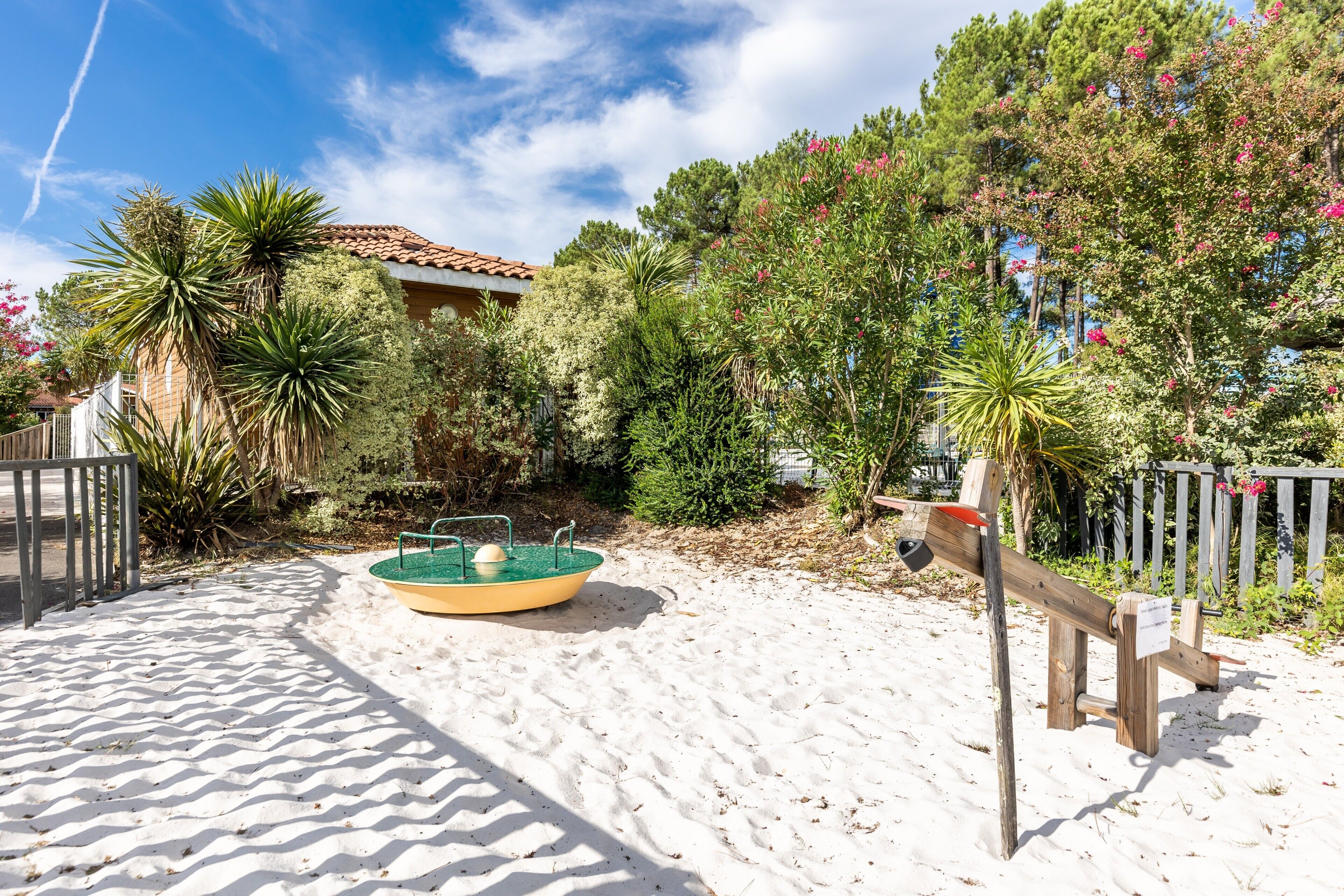 A sandy area with a small pool, surrounded by greenery and a wooden fence.