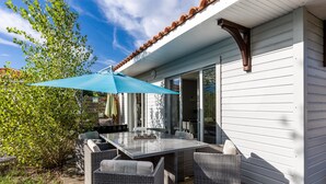 Un espacio de comedor al aire libre cubierto con mobiliario de mimbre y una mesa, contiguo a un edificio blanco con tejado de baldosas.