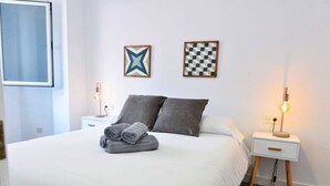 Una cama bien hecha con almohadas grises y toallas dobladas, dos cuadros en la pared, una mesita de noche con lámpara y un tocador también con lámpara.