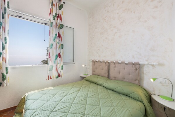 A bedroom with a bed, a window with curtains, a bedside lamp, and a wall-mounted mirror.