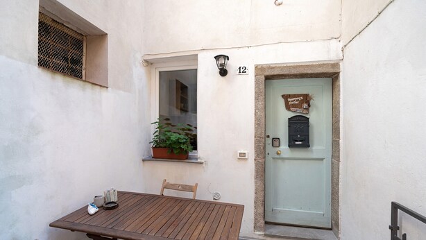 Une table et des chaises en bois sont placées à l’extérieur d’un bâtiment. La porte est marquée du numéro « 12 », et une enseigne se trouve au-dessus.