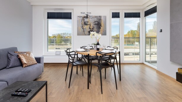 A modern dining area with a wooden table set for four, surrounded by black chairs. A grey sofa with patterned cushions is against the wall, and a black coffee table with a remote control is in the foreground. - Elegant Apartment in Gdansk | Parking | Close to the Beach (Gdansk)