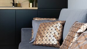 A modern living room with a dark sofa, geometric patterned cushions, a kitchen area with a sink and plants in the background. - Elegant Apartment in Gdansk | Parking | Close to the Beach (Gdansk)