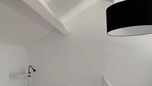 Une petite pièce blanche, avec un lit, un luminaire suspendu noir et une lampe blanche fixée au mur.
