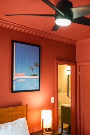 Una habitación con cama, ventilador de techo, un cuadro enmarcado y un baño visible a través de una puerta abierta.