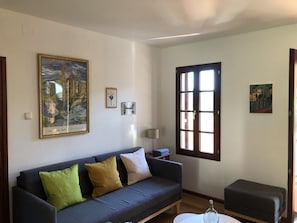Un soggiorno con un divano grigio scuro, cuscini verdi e gialli, un tavolino bianco e una bottiglia di vetro. Si notano tre quadri appesi alla parete, una finestra con persiane e una porta.