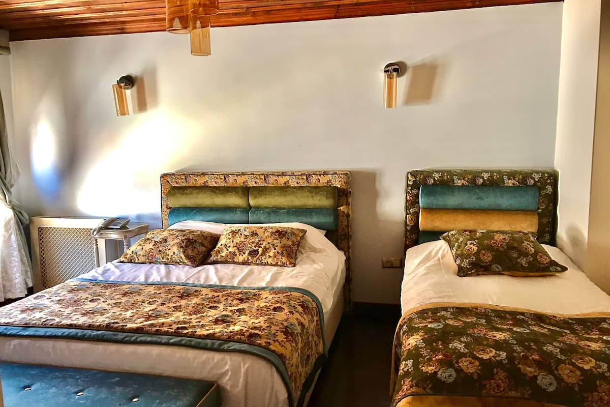 A room with two beds, each with floral-patterned bedding and headboards. There are bedside tables with lamps, and a window providing natural light.