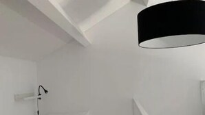 Et lille, hvidt rum med en seng, en sort hængende lampe og en hvid væglampe.