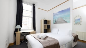 A hotel room with a bed, a desk, a chair, a window with curtains, a painting of a pier, and a radiator.