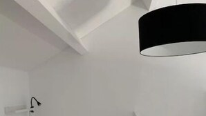 Une petite pièce blanche, avec un lit, un luminaire suspendu noir et une lampe blanche fixée au mur.
