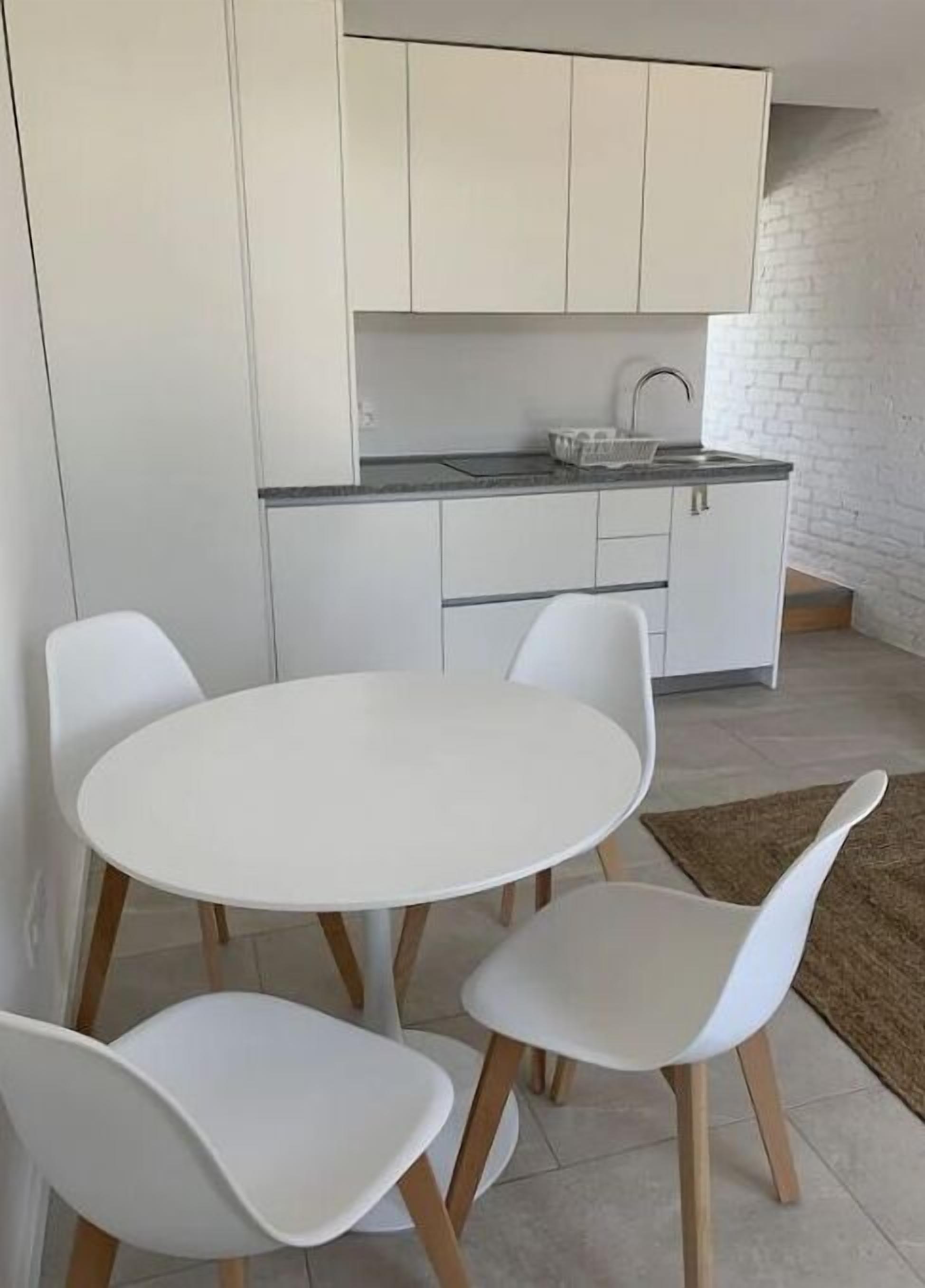 Una cocina compacta con gabinetes blancos, una encimera blanca y una mesa redonda con cuatro sillas blancas.